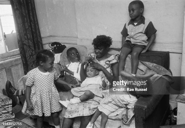 Woman sits combing the hair of a young child on a sofa, surrounded by other young boys and girls in her living room, in a property at a social...