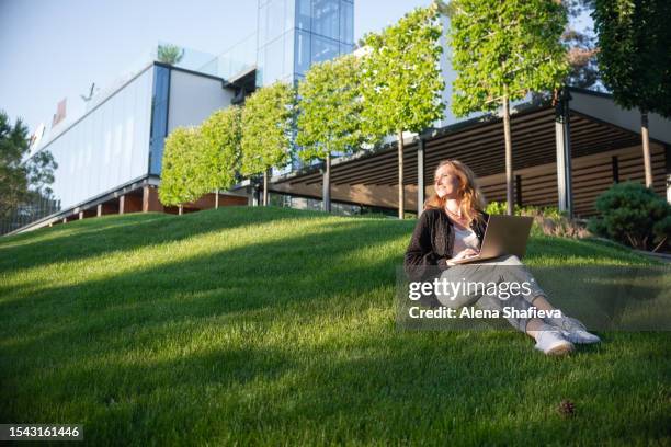 a young woman works on a laptop online among a green city park against the background of modern buildings office center - emerald city stock pictures, royalty-free photos & images