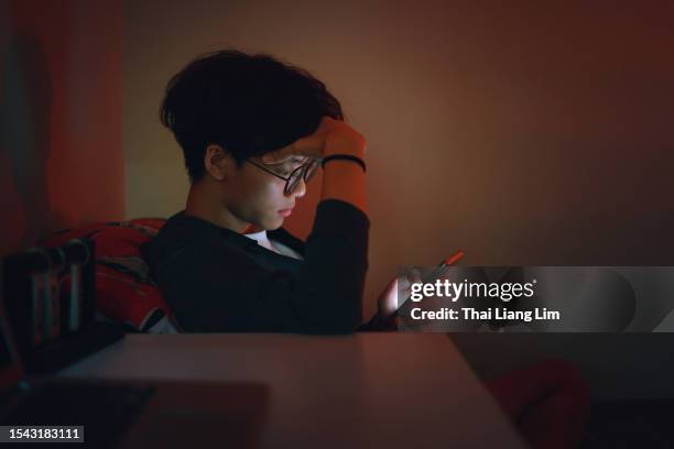 the upset teenage asian boy is reading alone on his smartphone late at night, caught in the grips of adolescent social media addiction - boys night stock pictures, royalty-free photos & images