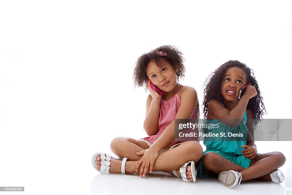 Studio shot of two girls (2-5, 6-7) sitting and talking on mobile phones