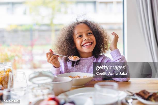 adorable black girl with curly hair smiling while eating breakfast in a bright kitchen - eating stock pictures, royalty-free photos & images