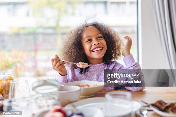 adorable black girl with curly hair smiling while eating breakfast in a bright kitchen - cereais de pequeno almoço imagens e fotografias de stock