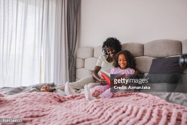 madre e hija negras felices disfrutando de su tiempo juntas y leyendo un cuento mientras están acostadas en una cama acogedora en un apartamento fresco - cama de matrimonio fotografías e imágenes de stock