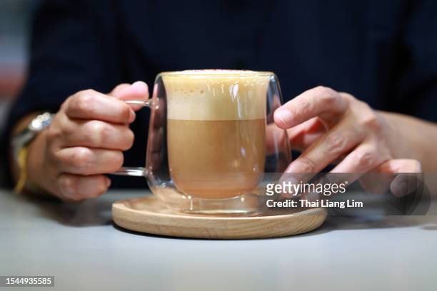 en un primer plano, las manos de una mujer sostienen delicadamente una taza de café con leche servido en una taza de vidrio de doble capa en un café. - capuccino fotografías e imágenes de stock