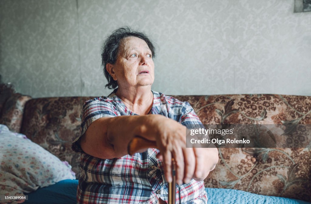 Poverty and disable old woman. Calm senior gray haired woman leaning on walking cane, sit on sofa in poor apartment