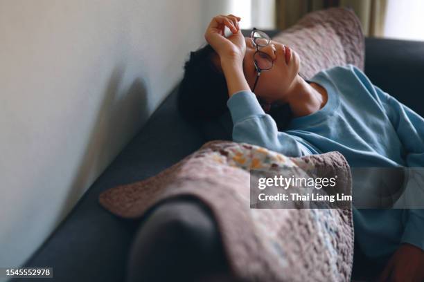 una adolescente asiática triste y deprimida sentada en el sofá de su casa con la mano en la frente - headache child fotografías e imágenes de stock