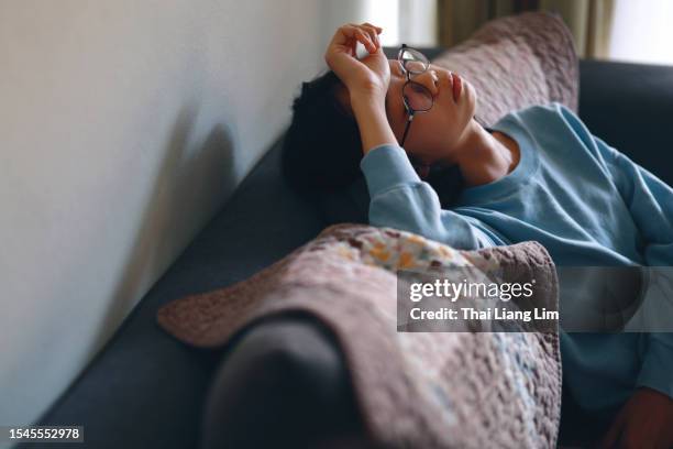 a sad and depressed asian teenage girl sitting on the sofa at home with her hand on her forehead - chinese girl stock pictures, royalty-free photos & images