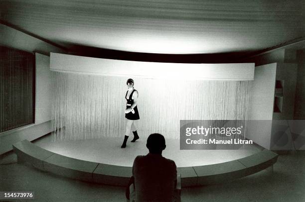 Andre COURREGES back sitting on a cushion in a square white ultra-modern living room watching a mannequin on a kind of little scene present a model...
