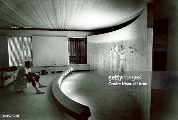 Andre COURREGES attitude in white shorts sitting on a cushion in a square white room looking at his ultra-modern models make a curtain of fibers...