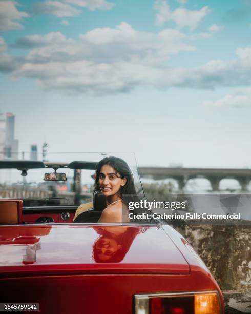 a woman glances over her shoulder as she sits in the back seat of a vintage, red convertible car. - vanuit een auto stockfoto's en -beelden