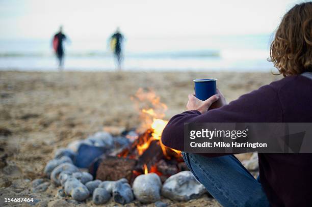 male surfer sitting by fire at beach party. - only mid adult men stock pictures, royalty-free photos & images