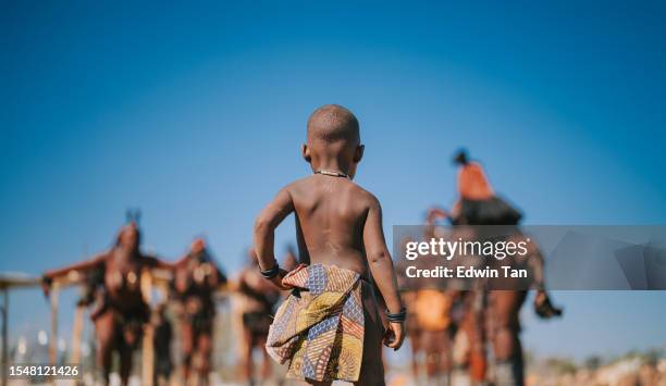 rückansicht namibianisches himba-kind, das dorfbewohner anschaut - afrikanischer volksstamm stock-fotos und bilder