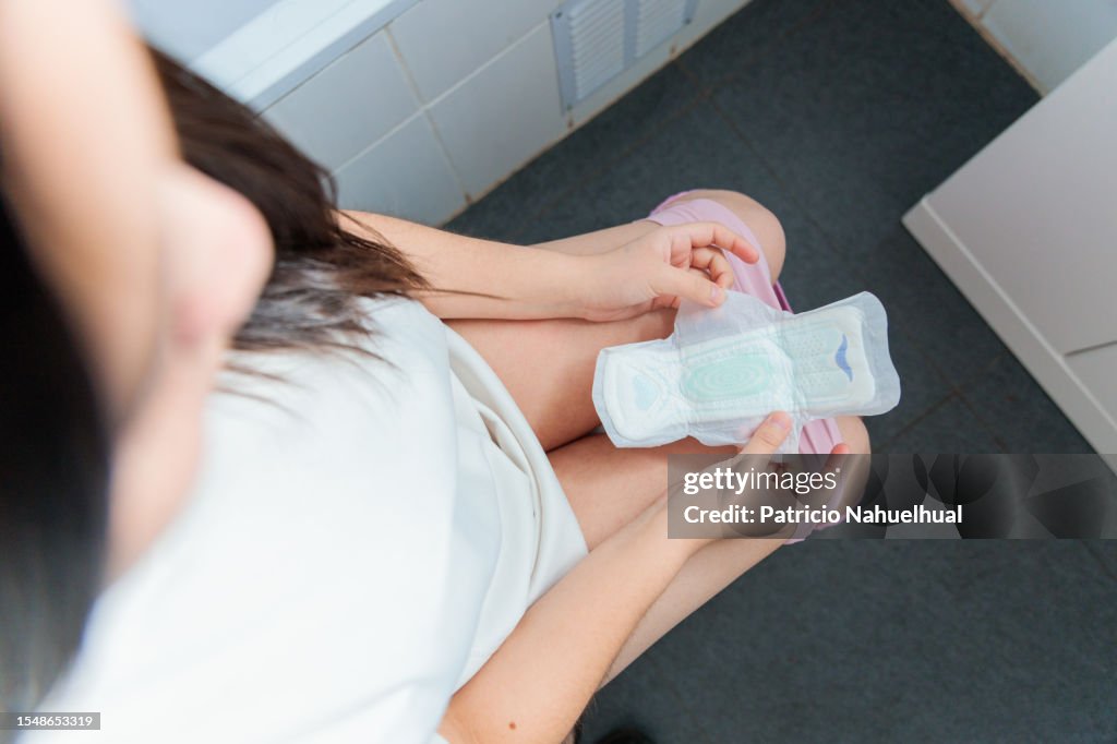 Young Woman, Dressed in a White Pajama, Seated in the Bathroom, Holding a Sanitary Napkin.