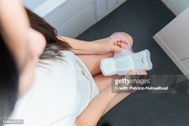 young woman, dressed in a white pajama, seated in the bathroom, holding a sanitary napkin. - damenbinde stock-fotos und bilder