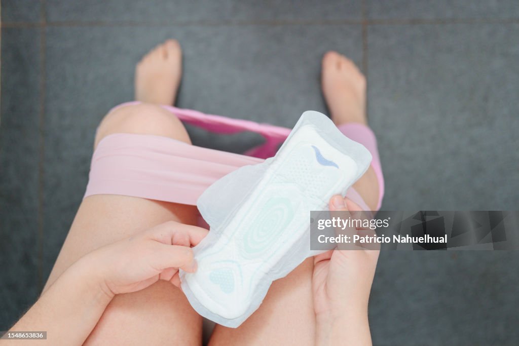 Unrecognizable Young Woman, Dressed in a White Pajama, Seated in the Bathroom, Holding a Sanitary Napkin.