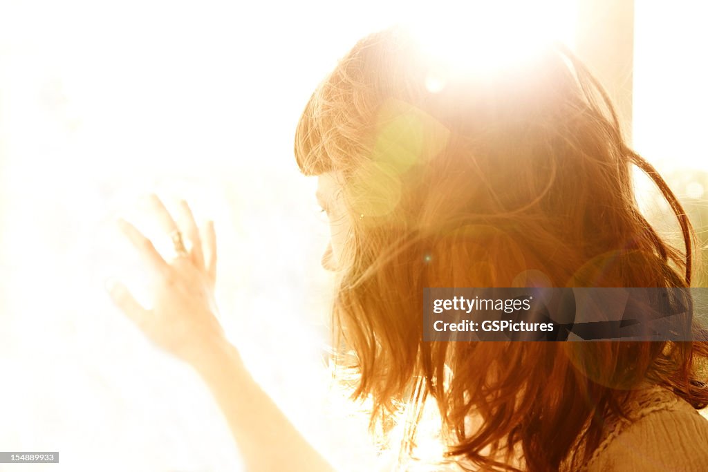 Redhead woman backlit by sun with hand on glass window