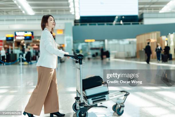 young asian business woman walking in airport terminal with luggage trolley - luggage trolley stock pictures, royalty-free photos & images