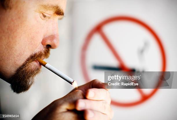 man lighting cigarette in front of no smoking sign - emphysema stock pictures, royalty-free photos & images