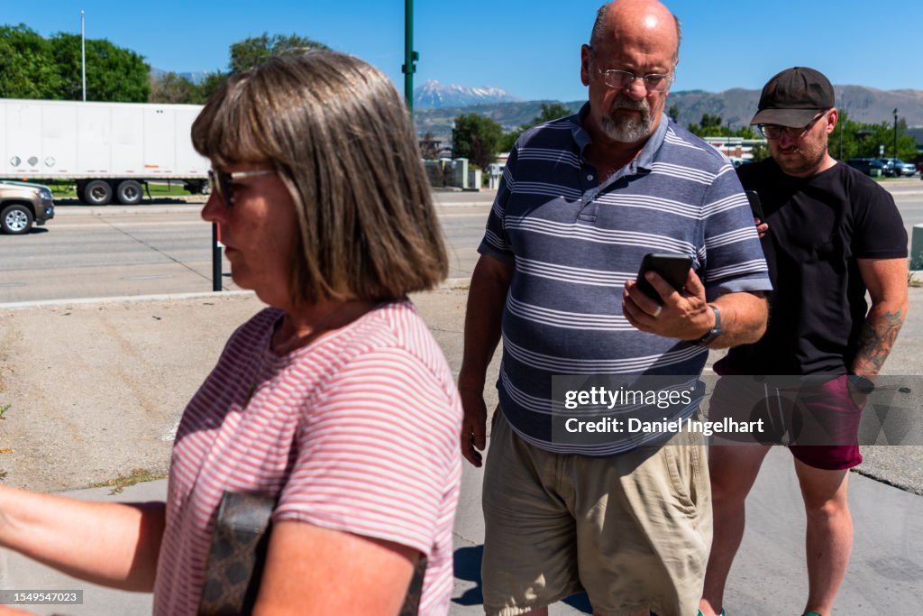 In line at sunny ATM: Woman (50s) in pink shirt, jeans; early 30s man (black hat, tattoos); mid-50s man (blue/white striped shirt, cargo shorts).