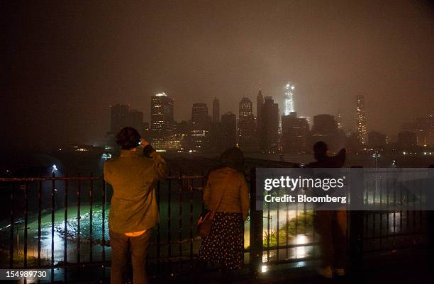 Onlookers watch while flood waters cover a promenade in Brooklyn as numerous buildings in Lower Manhattan, background, stand in darkness in New York,...