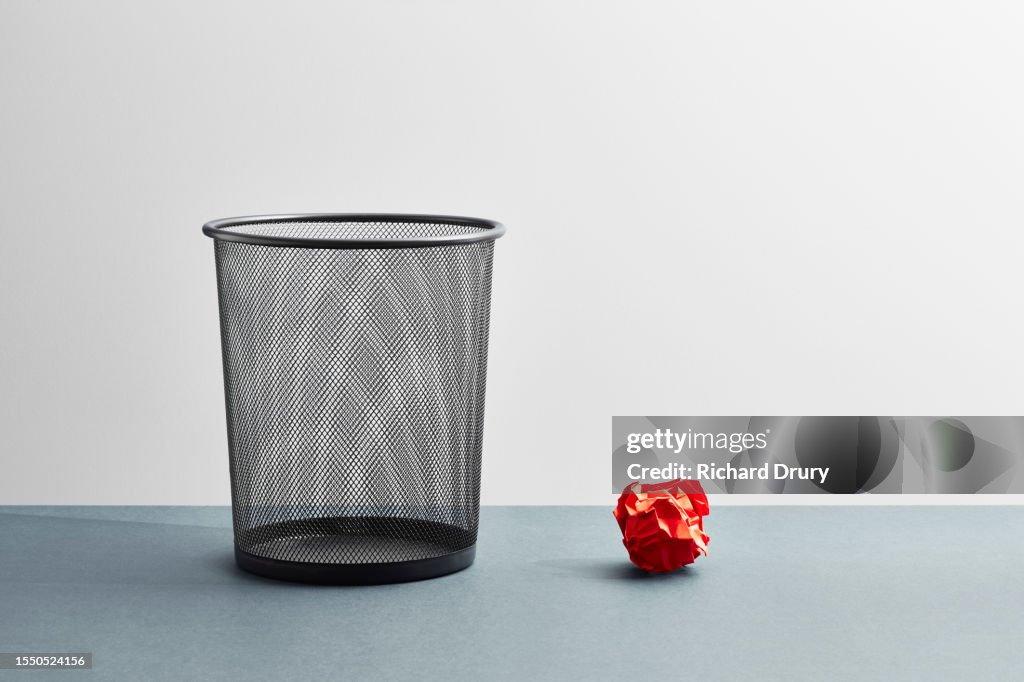 An empty wastepaper basket with one red crumpled paper ball beside it