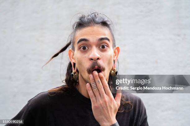 young multiracial man with long hair, dreadlocks, nose rings, piercings covering his mouth with an oops gesture, whoops, error, mistake, speak no evil - intim piercing bildbanksfoton och bilder