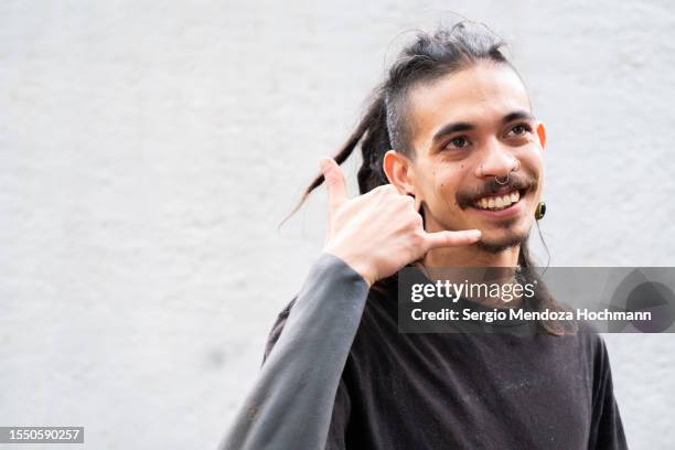 young multiracial man with long hair, dreadlocks, nose rings, piercings and sleeve tattoos making a "call me on the phone" hand gesture - call me stock pictures, royalty-free photos & images