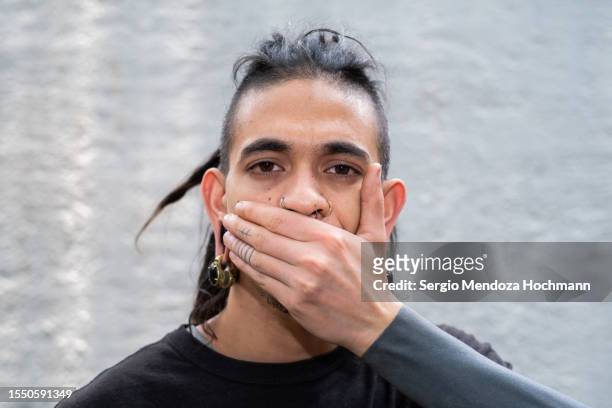 young multiracial man with long hair, dreadlocks, nose rings, piercings and sleeve tattoos looking at the camera and covering his mouth, speak no evil - intim piercing bildbanksfoton och bilder