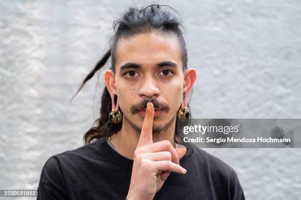 young multiracial man with long hair, dreadlocks, nose rings, piercings looking at the camera with a finger on his lips, asking for quiet, silence - man finger on lips stock pictures, royalty-free photos & images