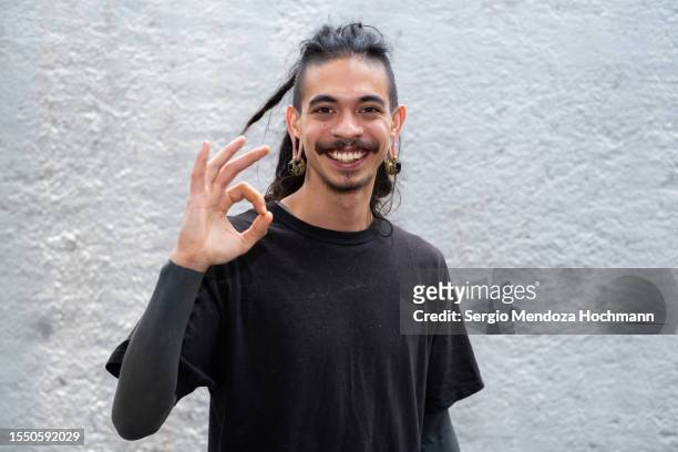 young multiracial man with long hair, dreadlocks, nose rings, piercings and sleeve tattoos looking at the camera and giving the ok sign - mh stock pictures, royalty-free photos & images