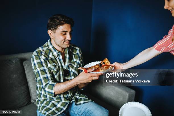 cheerful gen z woman giving pizza slice to male friend sitting on couch - pizza temptation stock pictures, royalty-free photos & images