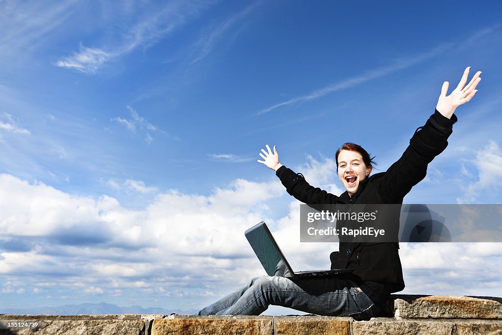 Happy young woman with laptop on cliffside wall