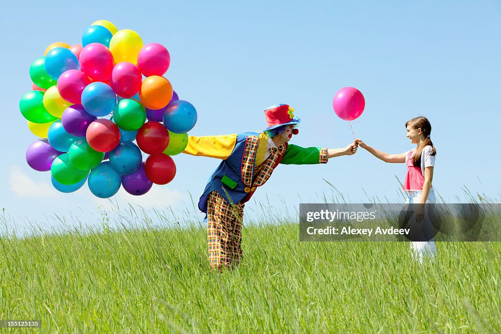 Clown with balloon bouquet giving one to a girl in a field