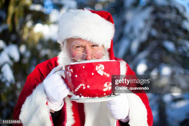 cheerful santa claus sipping cocoa from oversized mug in snow - slagroom stockfoto's en -beelden