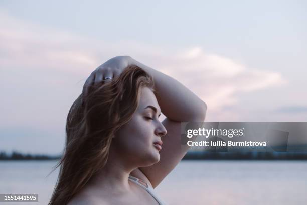 attractive young woman with long blond hair relishing peaceful ambiance of lake beach at dusk - chubby face stockfoto's en -beelden