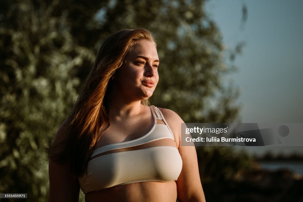 Serene woman with long red hair standing on river beach in flattering bikini