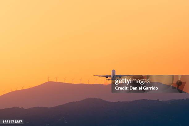 golden takeoff towards a sustainable future, airplane soaring at sunset with mountains and renewable energy wind turbines in the background - launch stock pictures, royalty-free photos & images
