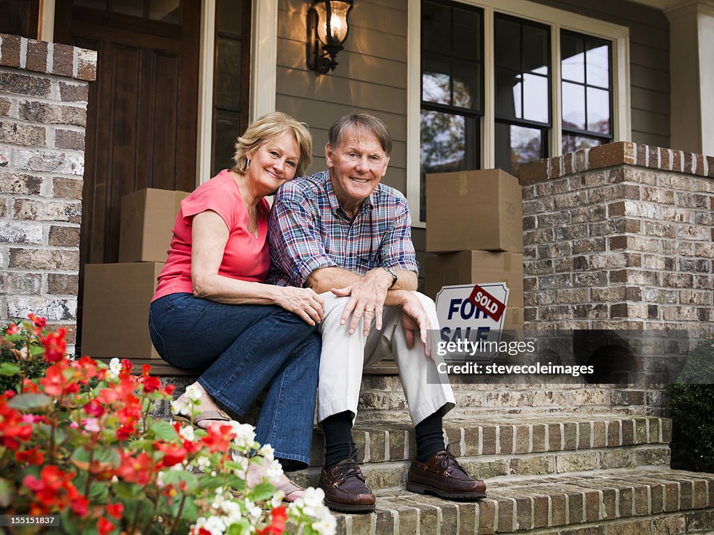 Mature Couple Sitting Outside House With For Sale Sign