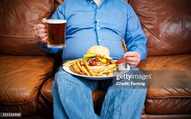 hombre con sobrepeso de comer en un sofá en la sala de estar - barrigón fotografías e imágenes de stock