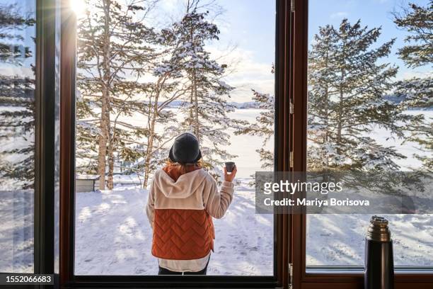 a woman in winter clothes stands outside the window drinking tea from a thermos in awe of the winter countryside. - sunlight window stock pictures, royalty-free photos & images