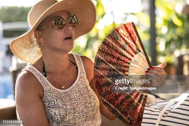 woman suffering a heat wave using a fan, sitting on a couch in the restaurant terrace. - ac weary stockfoto's en -beelden