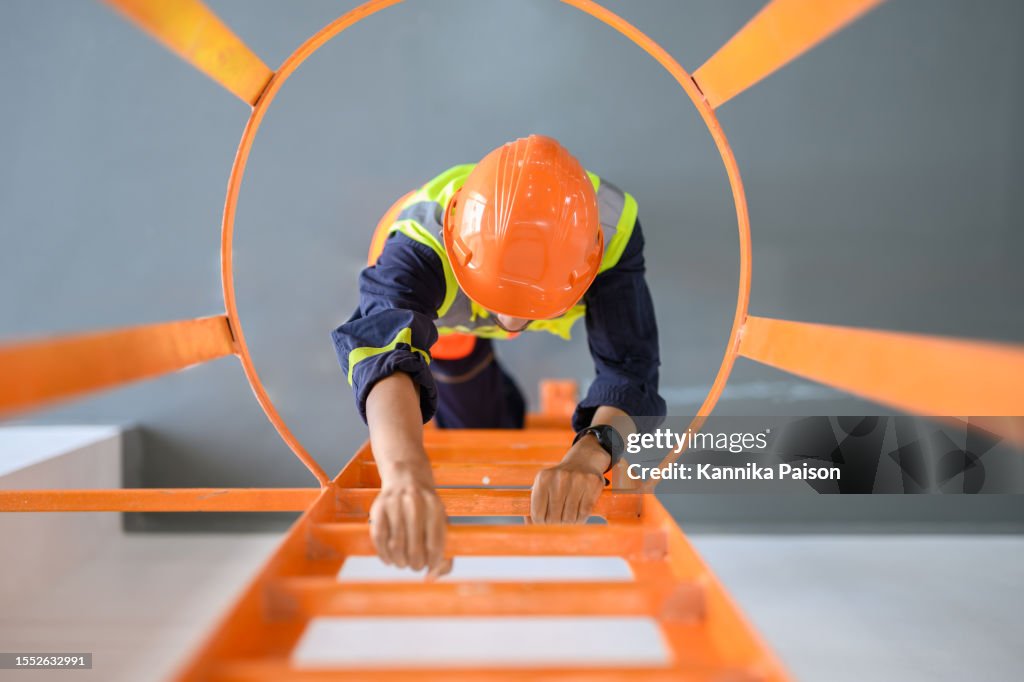 Vista dall'alto angolo di vista verso il basso su giovane tecnico asiatico che sale la scala per ispezionare o manutenere la macchina nella sottostazione di trasformazione elettrica ad alta tensione. Concetto di lavoratore di occupazione industriale.