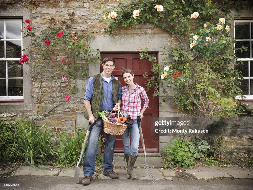 Portrait of farming couple with basket of organic vegetables and gardening tools standing in front of farmhouse doorway