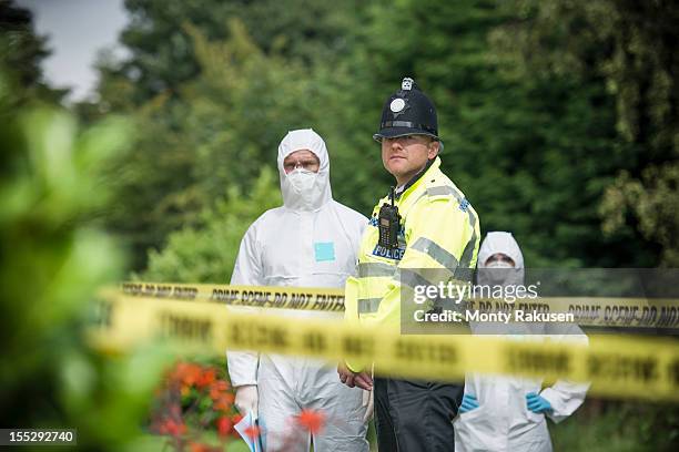 policeman and forensic scientists at crime scene with police tape in foreground - scena del crimine foto e immagini stock