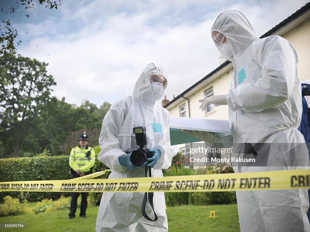 Forensic scientists in discussion at crime scene, one holding camera, policeman in background