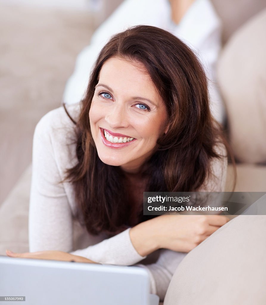 Smiling mature lady lying on couch and using laptop
