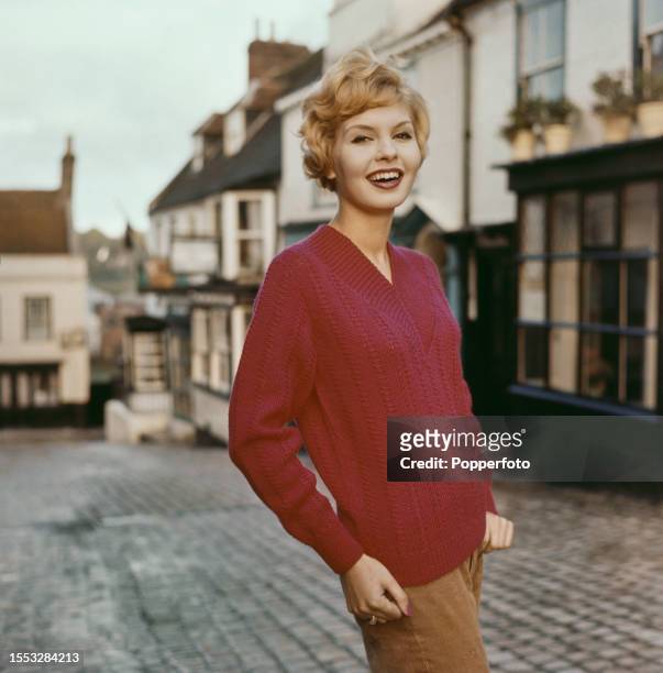 Exterior view of a female fashion model posed wearing a red cable knit jersey sweater with a V-neck, she stands on a cobbled street in an English...