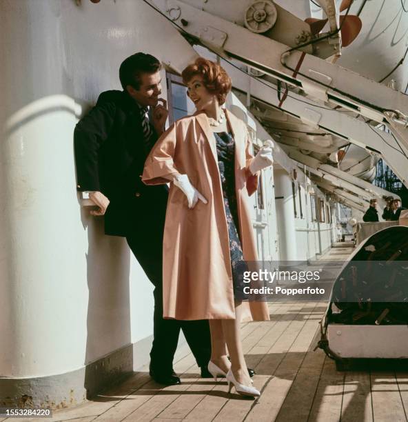 Female fashion model posed wearing an apricot duster coat over a blue dress with pink flower pattern, she stands with a man on the passenger...