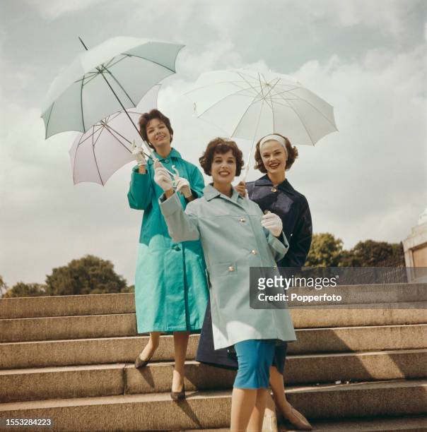 Three female fashion models posed wearing macs and rainwear in dark blue, light blue and turquoise, they hold umbrellas and stand on the steps of the...