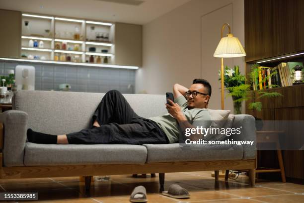 asiatischer mann mittleren alters, der auf dem sofa liegt und auf sein telefon schaut - handsome middle eastern men stock-fotos und bilder